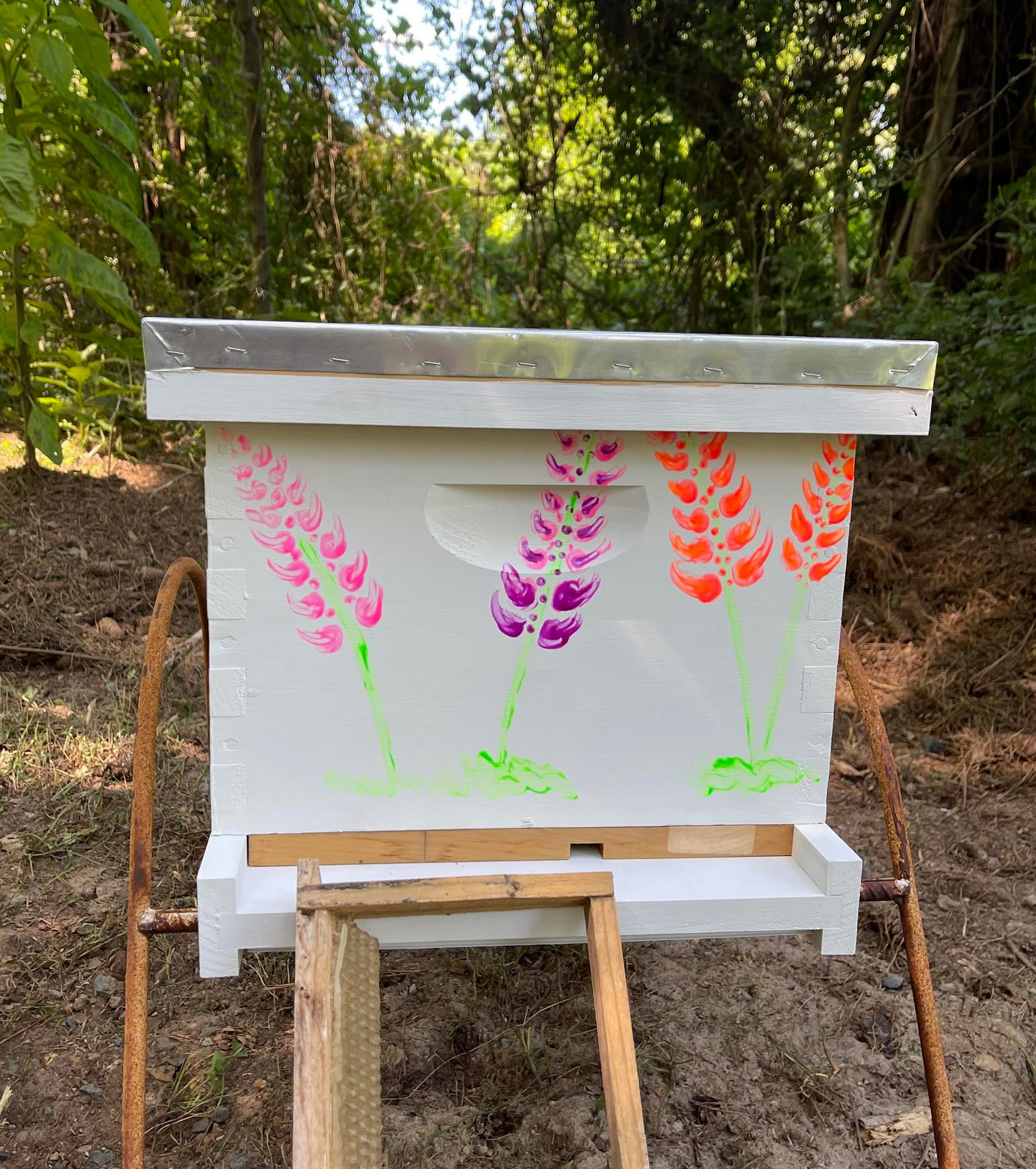 A white beehive with a galvanized telescoping cover, painted on the front with three lupine flower stalks in pink, purple, and orange, on a metal and wood stand in a wooded clearing