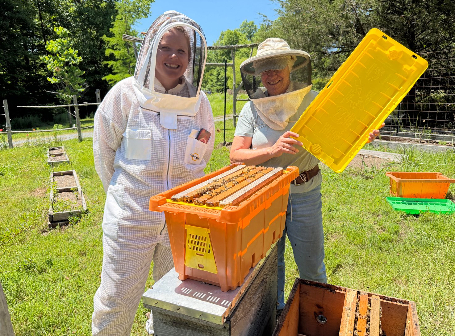 Lilly-Gray Warren in a white beekeeping suit and Kory Goldsmith holding the inner cover of a Pro Nuc, with frames of bees visible